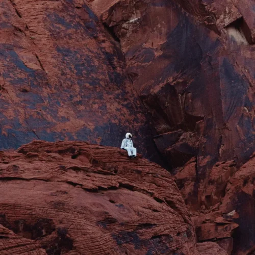 shopthietke com An astronaut in a white spacesuit sitting on red rocky terrain of a canyon with high cliffs in the background