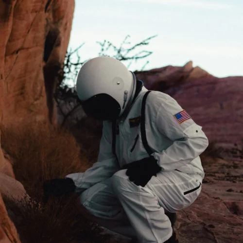 shopthietke com Astronaut in a white space suit kneeling next to red rock formations in a desert landscape with mountains in the background