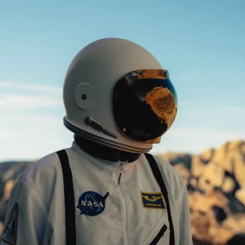 shopthietke com Person in an astronaut suit with a helmet reflecting a rocky landscape standing outdoors with mountains in the background